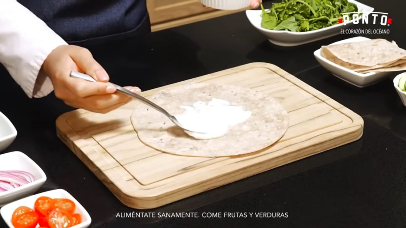 Prepara la base del wrap: Coloca la tortilla integral sobre una superficie plana y agrega una capa de yogurt griego sin azúcar. Esto le dará muchísima cremosidad y un sabor delicioso al wrap. Si prefieres una opción más fresca, también puedes añadirle un poco de limón.